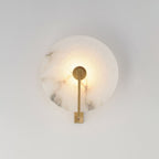 Close view of this Clérachic wall sconce in brushed brass, its round alabaster disc lit warm on a grey wall.