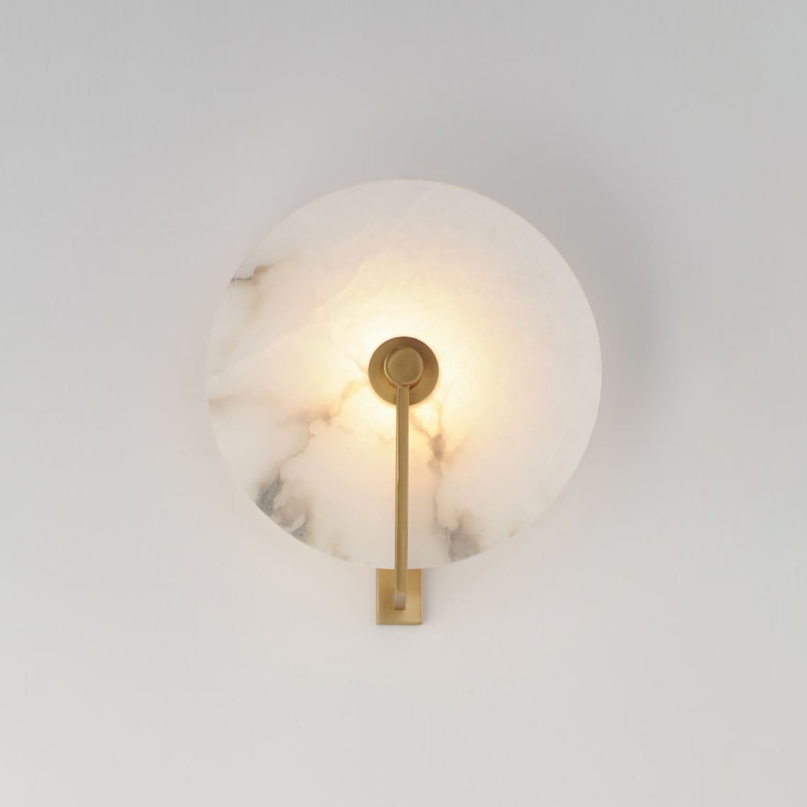 Close view of this Clérachic wall sconce in brushed brass, its round alabaster disc lit warm on a grey wall.
