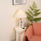 Clérachic table lamp lit with its cord reaching a wall socket, framed print above and fern fronds near the shade.