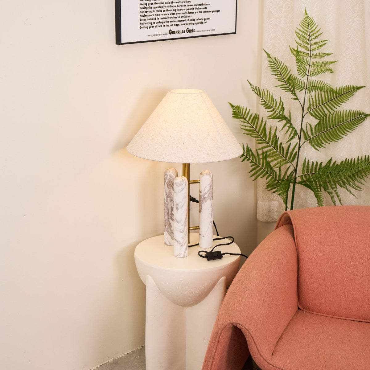 Clérachic table lamp lit with its cord reaching a wall socket, framed print above and fern fronds near the shade.