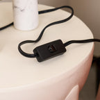 Close-up of one Clérachic lamp column's marble base and its black inline cord switch on a pale pink table edge.