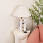 Clérachic table lamp with its cord hanging past a large fern frond, the coral armchair visible just below.
