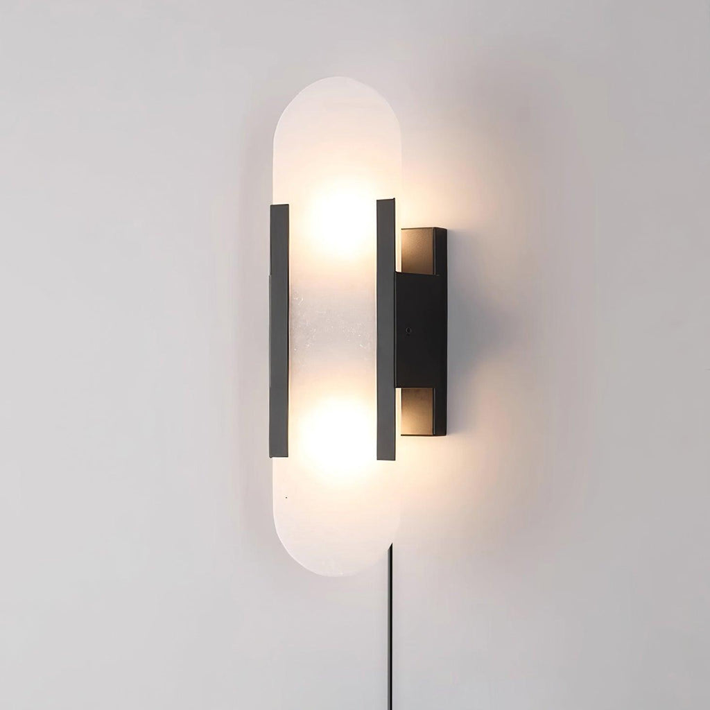 Black-finish Kristy alabaster wall lamp lit on a pale wall, its plug-in power cord running down below