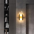 Black and brass Kristy sconce lit on a dark concrete wall beside a tall shelf of books
