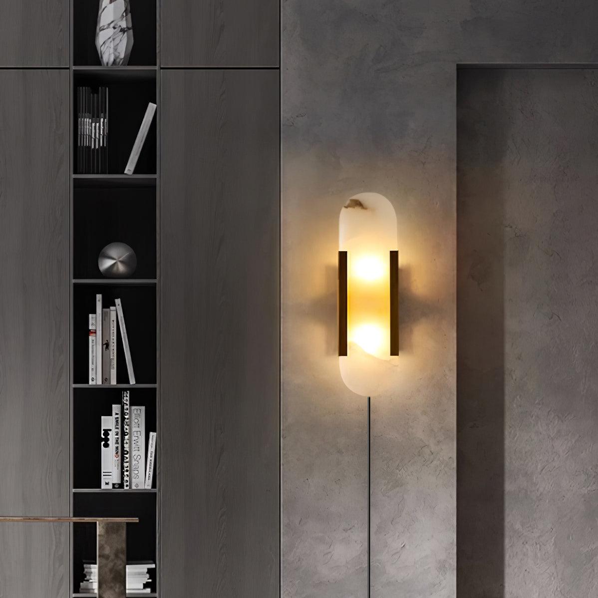 Black and brass Kristy sconce lit on a dark concrete wall beside a tall shelf of books