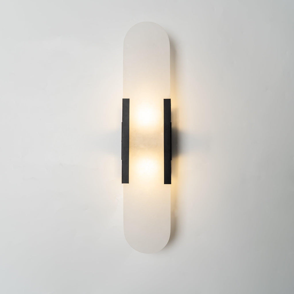 Clérachic elongated black-finish Olraeia sconce head-on, warm light spilling either side onto a plaster wall