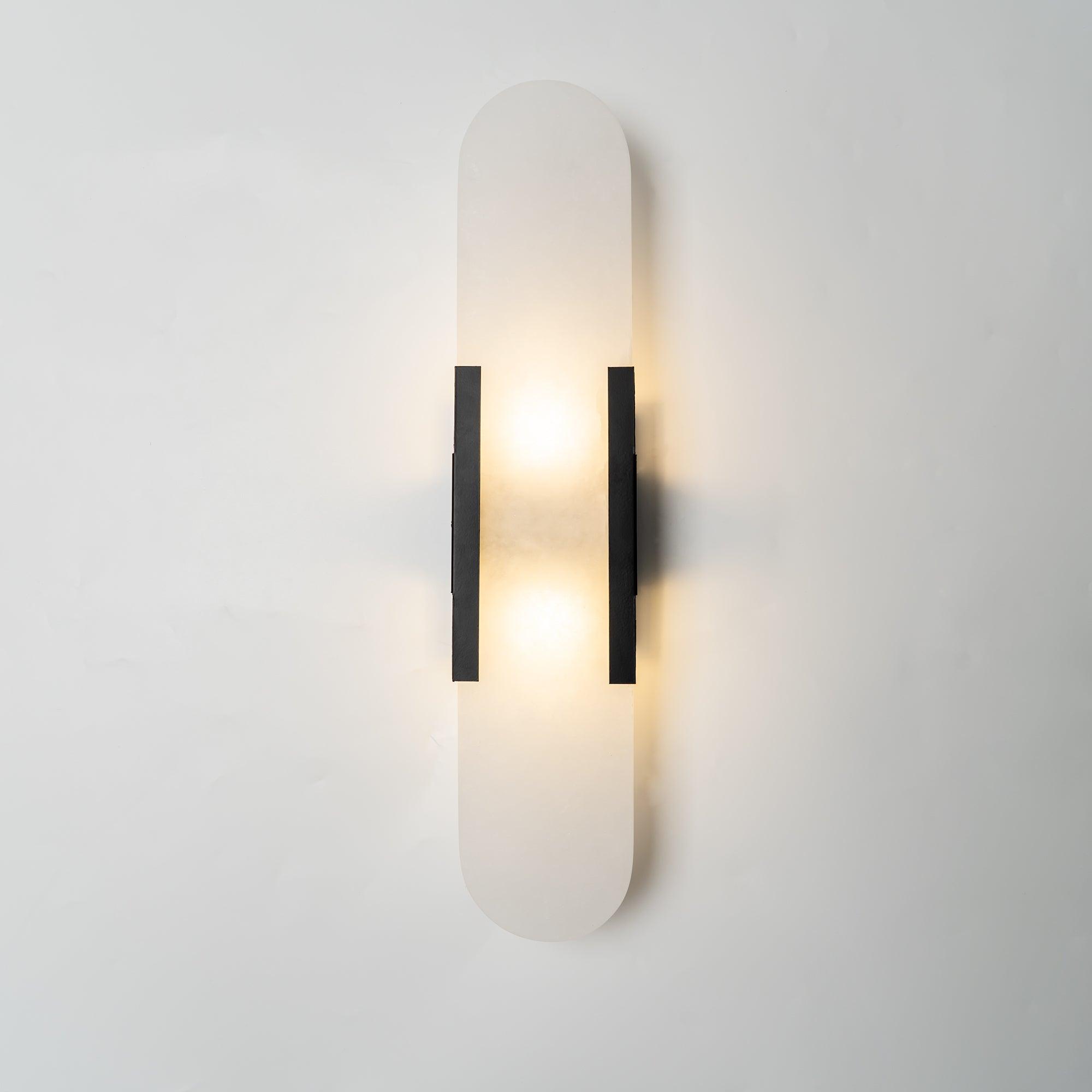 Clérachic black Olraeia sconce lit on a pale plaster wall, the stone panel's soft cloudy grain showing through