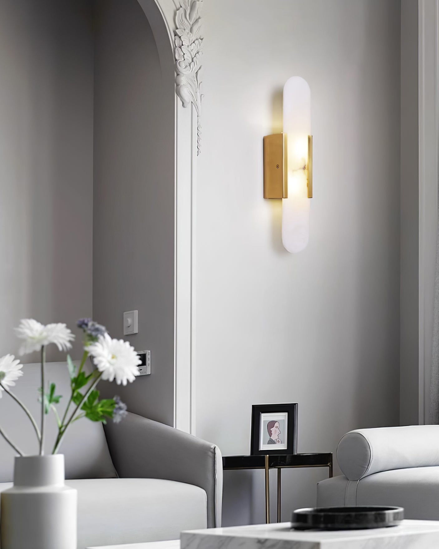 Clérachic olraeia sconce lit beside an ornate plaster arch in a pale living room with grey sofas and daisies