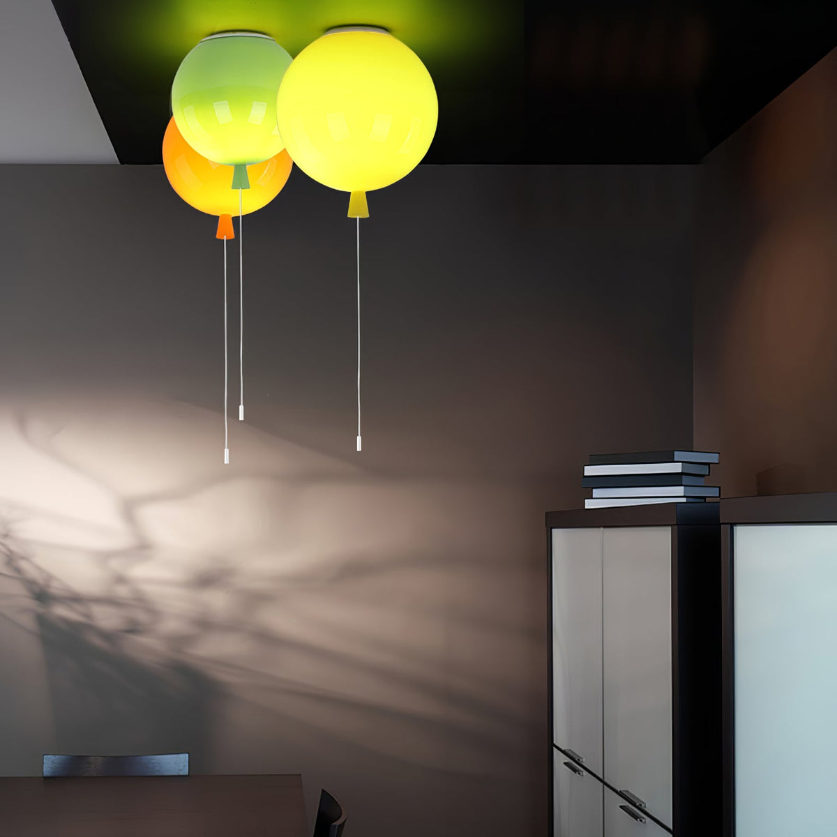 Casting long shadows in a dark room, three Clérachic balloon lights in green, orange and yellow hang near a bookshelf.
