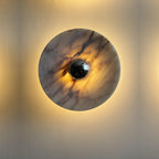 Macro close-up of the Clérachic marble disc wall light, its black centre knob ringed by a strong warm amber glow on a dark wall.