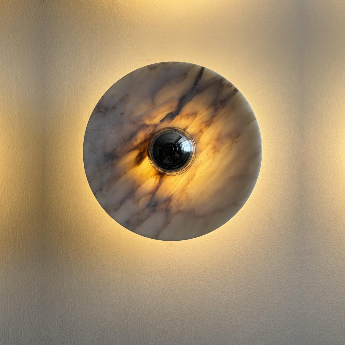 Macro close-up of the Clérachic marble disc wall light, its black centre knob ringed by a strong warm amber glow on a dark wall.