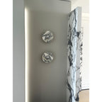 Two unlit Clérachic marble wall lights hang vertically beside a tall mirror and a veined marble slab in a daylit hallway.
