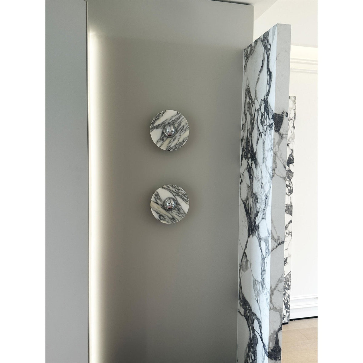 Two unlit Clérachic marble wall lights hang vertically beside a tall mirror and a veined marble slab in a daylit hallway.