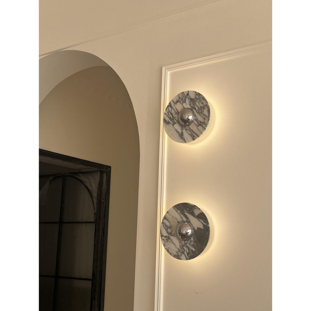 Two Clérachic marble wall lights glow warm amber in a corner beside a bright glazed door and a floor-length curtain.