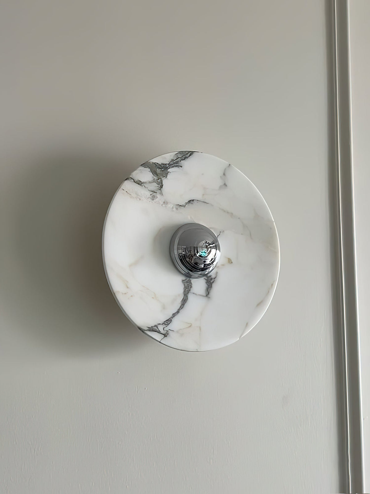 A single Clérachic marble wall light with a chrome centre is mounted, unlit, on a plain wall beside a door frame edge.