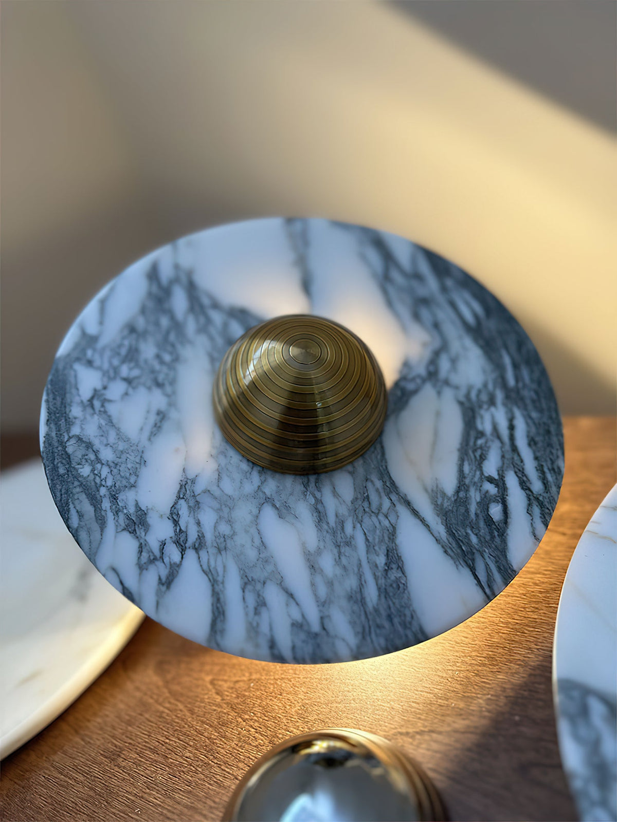 A Clérachic marble disc tilts on a dresser corner, its brass dome centre lit by raking sunlight, a chrome dome partly visible below.