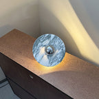 A Clérachic marble disc with a chrome centre rests propped on a wooden dresser corner, warm light glowing beneath its edge.