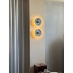 Two Clérachic marble wall lights glow warm amber, stacked vertically in a curtained bedroom corner beside a dark plinth.