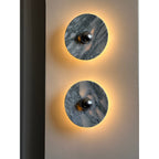 Close crop of two Clérachic marble wall lights mounted vertically on a plain wall pillar, both glowing strongly amber.