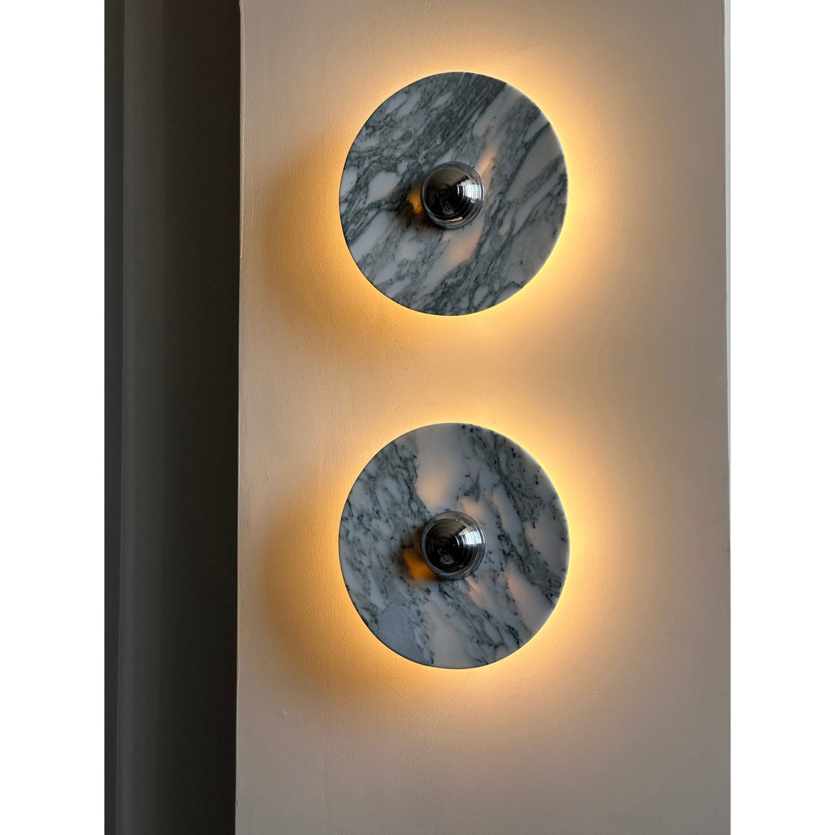 Close crop of two Clérachic marble wall lights mounted vertically on a plain wall pillar, both glowing strongly amber.