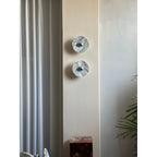 A distant view of two Clérachic marble wall lights, unlit, beside a curtain and a dark marble plinth topped with a small object.