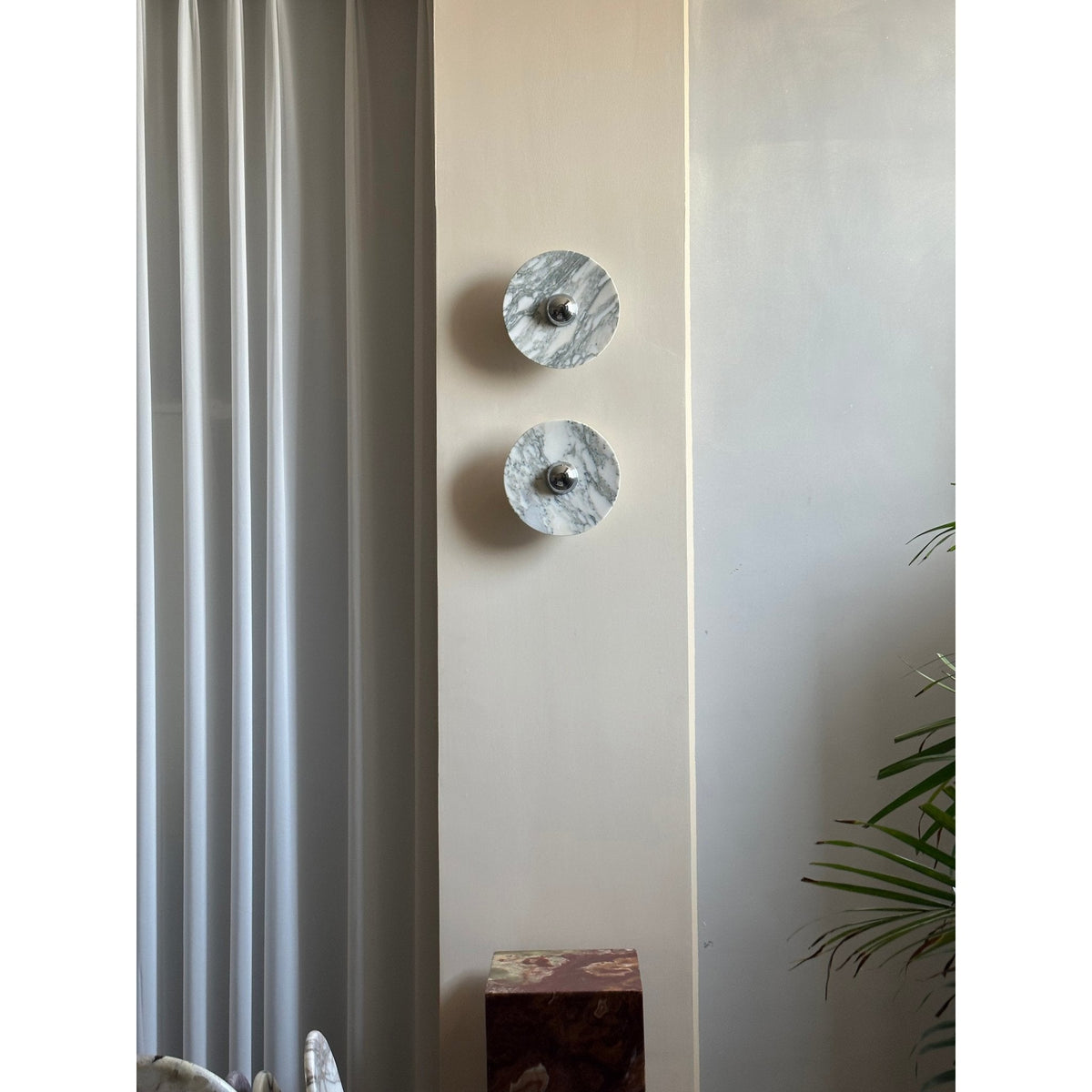 A distant view of two Clérachic marble wall lights, unlit, beside a curtain and a dark marble plinth topped with a small object.