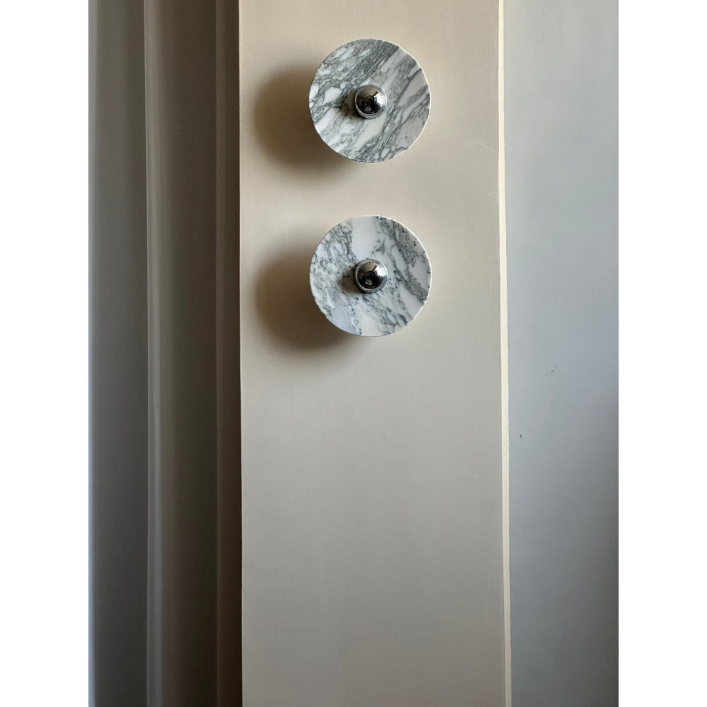 Two unlit Clérachic marble wall lights mounted vertically on a full-height wall pillar, seen in daylight beside a curtain.