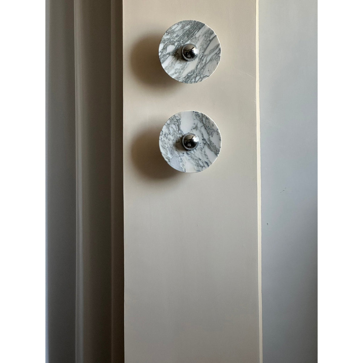 Two unlit Clérachic marble wall lights mounted vertically on a full-height wall pillar, seen in daylight beside a curtain.