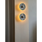 Two Clérachic marble wall lights glow warm amber on a wall pillar, photographed from a medium distance beside a curtain.