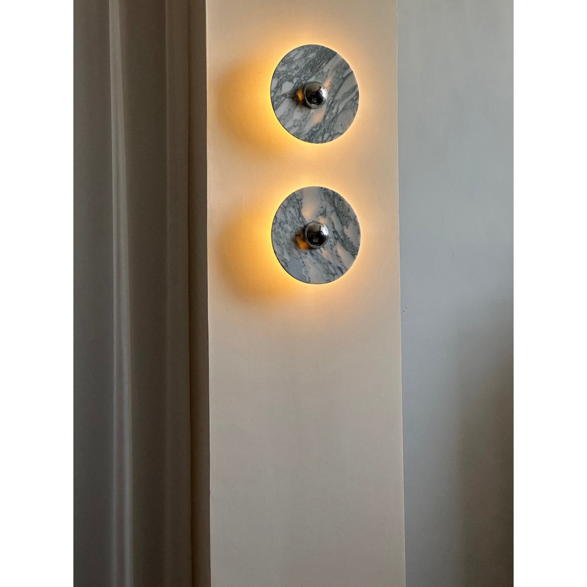 Two Clérachic marble wall lights glow warm amber on a wall pillar, photographed from a medium distance beside a curtain.
