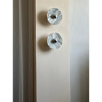 Two unlit Clérachic marble wall lights mounted vertically, photographed close-up in daylight beside a curtained pillar.