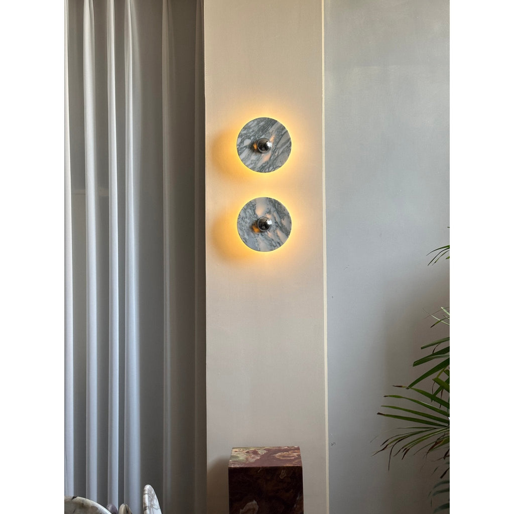 A smaller, distant crop of two glowing Clérachic marble wall lights beside a curtain, dark plinth and trailing plant leaves.
