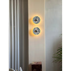 A smaller, distant crop of two glowing Clérachic marble wall lights beside a curtain, dark plinth and trailing plant leaves.