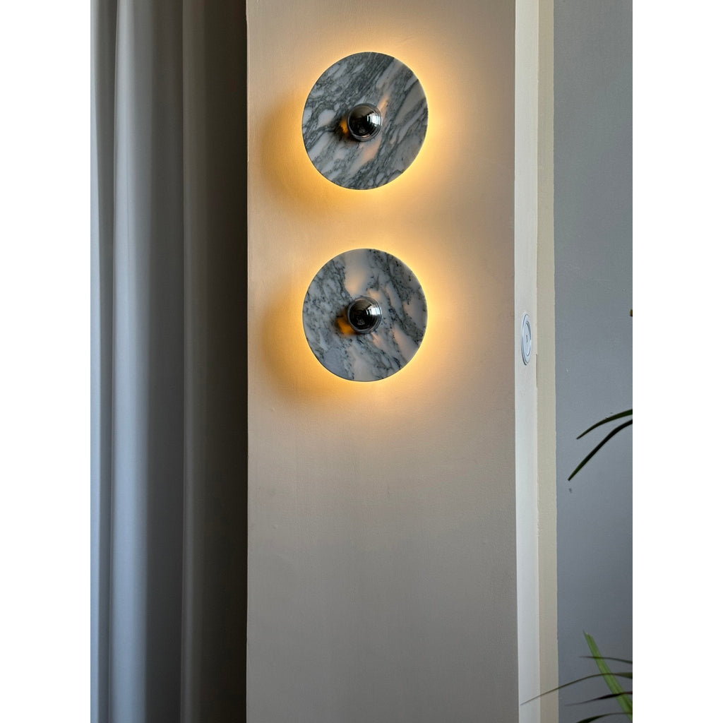 Two Clérachic marble wall lights glow warm amber on a wall pillar, with a light switch plate visible beside the lower fixture.