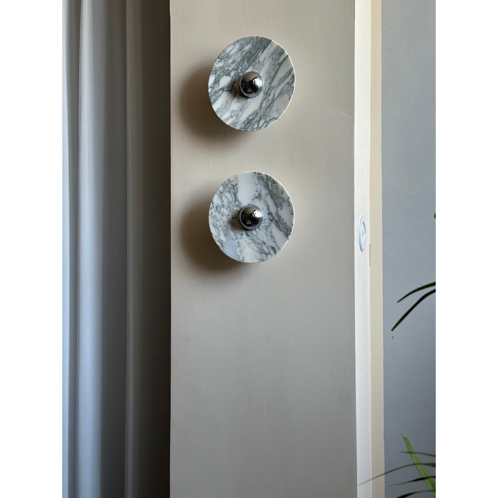 A distant, unlit view of two Clérachic marble wall lights beside a curtain and door frame, with a trailing plant leaf in shot.