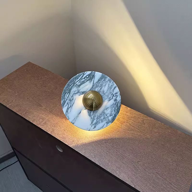 A grey-veined Clérachic marble disc with a brass centre knob is propped on a wooden dresser corner, lit warm from below.