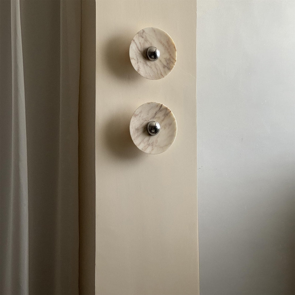 Two cream marble Clérachic wall lights, softly lit, are mounted vertically in a narrow wall nook beside a curtained window.