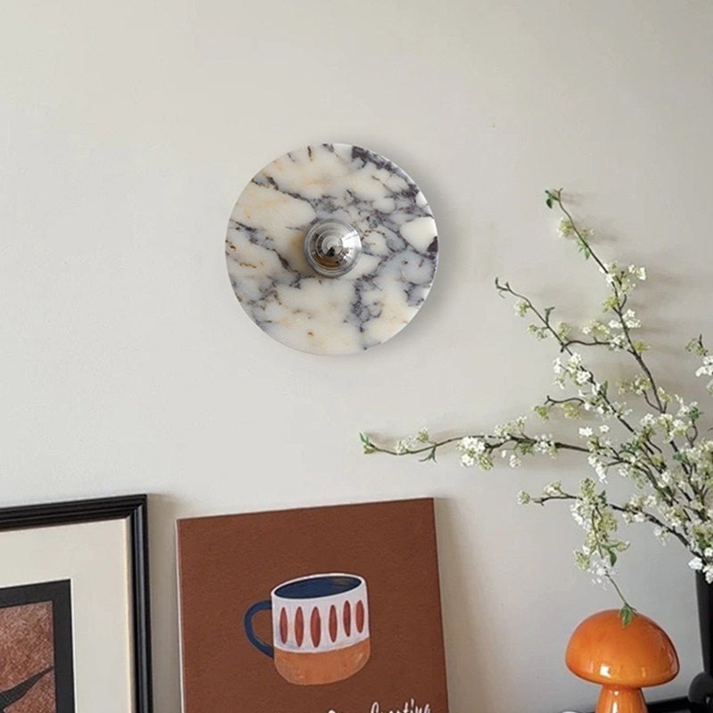 A grey-veined Clérachic marble wall light hangs above a framed print and a vase of blossom branches in the same entryway corner.