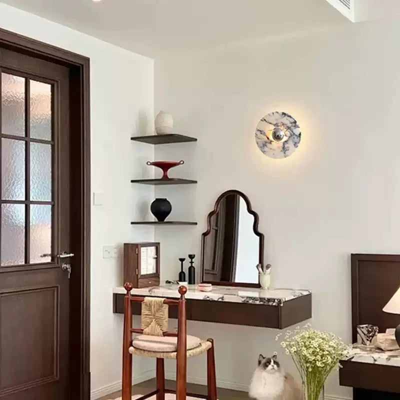 A Clérachic marble wall light glows above a dark wood dressing table in an entryway, with a cat resting nearby and blossoms in a vase.