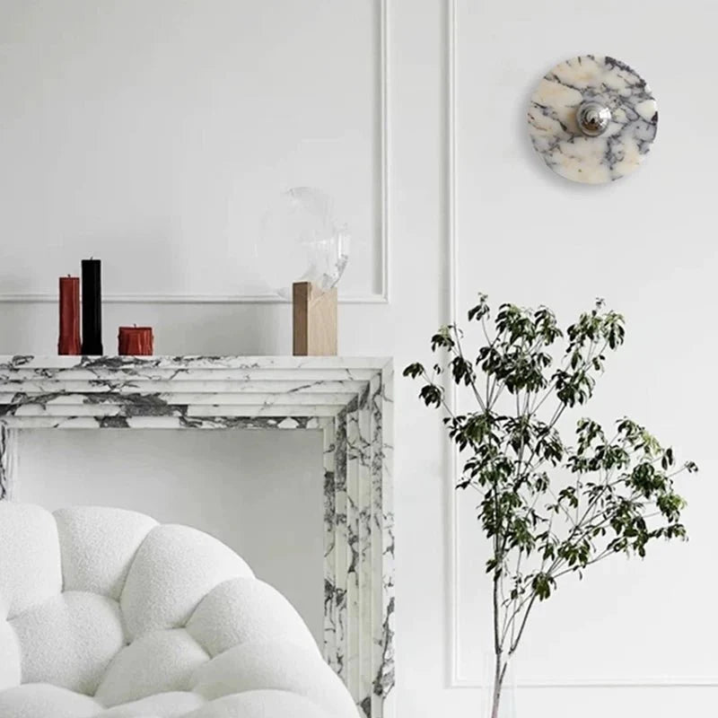 A tall, narrow crop frames the unlit Clérachic wall light above the fireplace mantel, with the candle holders and chair edge below.