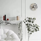 An unlit Clérachic marble wall light hangs above a white marble fireplace, beside slim candle holders and a bouclé armchair.
