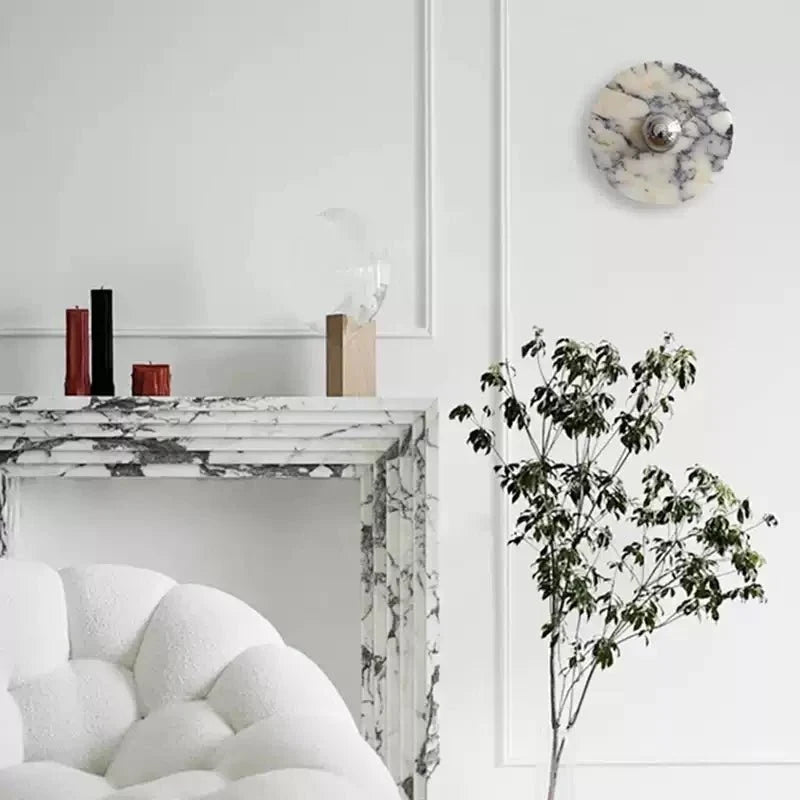 An unlit Clérachic marble wall light hangs above a white marble fireplace, beside slim candle holders and a bouclé armchair.