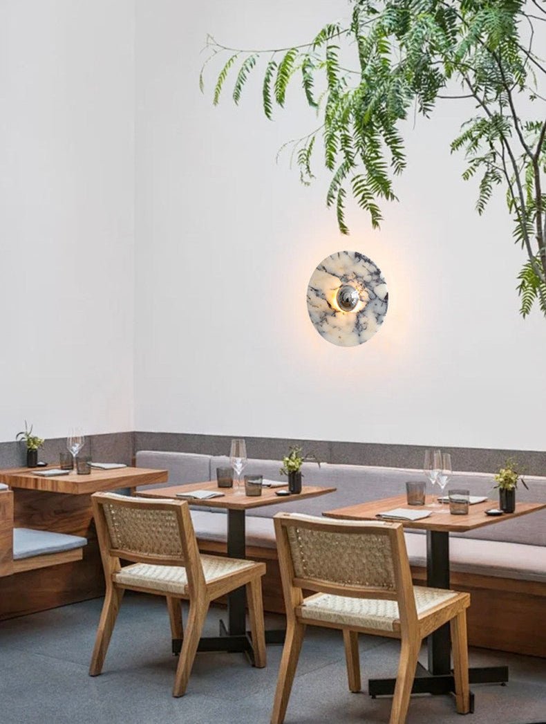 A Clérachic marble wall light glows warm amber beside wooden banquette seating in a restaurant dining nook with hanging greenery.