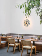 A Clérachic marble wall light glows warm amber beside wooden banquette seating in a restaurant dining nook with hanging greenery.