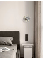 A single unlit Clérachic marble wall light is mounted in a bedroom corner beside a dark headboard and a pale pedestal table.
