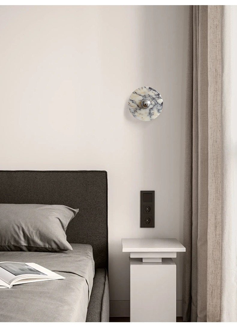 A single unlit Clérachic marble wall light is mounted in a bedroom corner beside a dark headboard and a pale pedestal table.