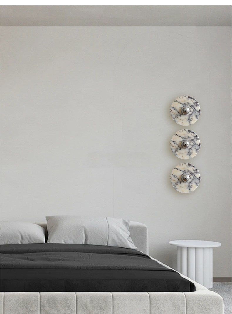 Three Clérachic marble wall lights are stacked vertically in a bedroom corner, all glowing warm amber beside a round side table.