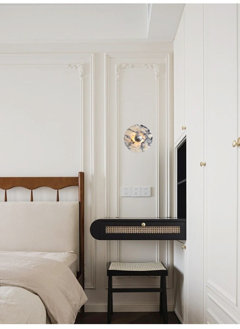 A Clérachic wall light glows warm amber above a black cane bench and stool beside a wood bed frame and panelled wardrobe doors.