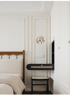 A Clérachic wall light glows warm amber above a black cane bench and stool beside a wood bed frame and panelled wardrobe doors.