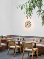 A wider dining-room crop of the glowing Clérachic wall light beside banquette seating and trailing greenery overhead.